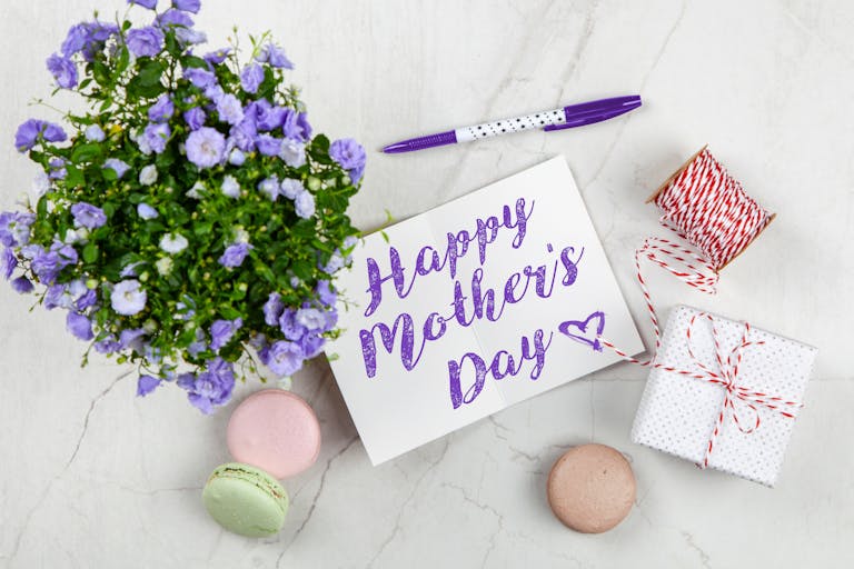 Flat lay of Mother's Day card with purple flowers, gift, macarons, and a pen indoors.