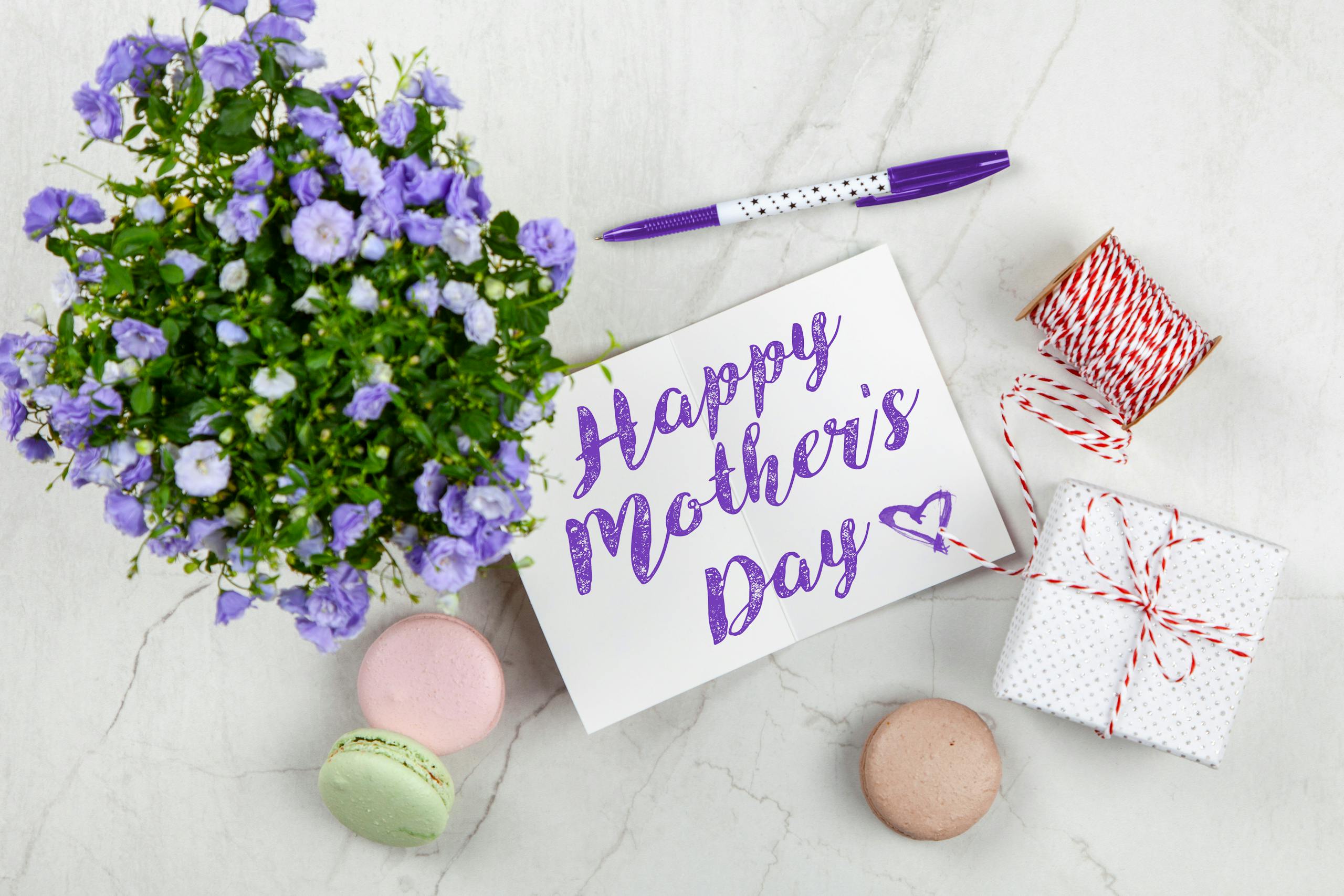 Flat lay of Mother's Day card with purple flowers, gift, macarons, and a pen indoors.
